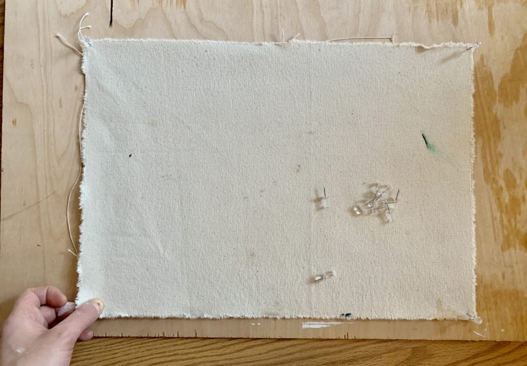 A hand holds a corner of a piece of canvas while pinning it to a wood panel